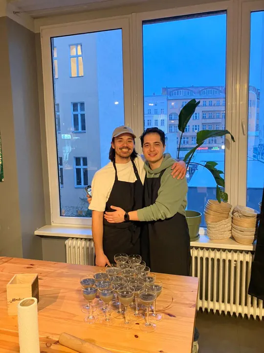 Zwei Kollegen in Schürzen umarmen sich hinter einem Tisch mit gefüllten Gläsern