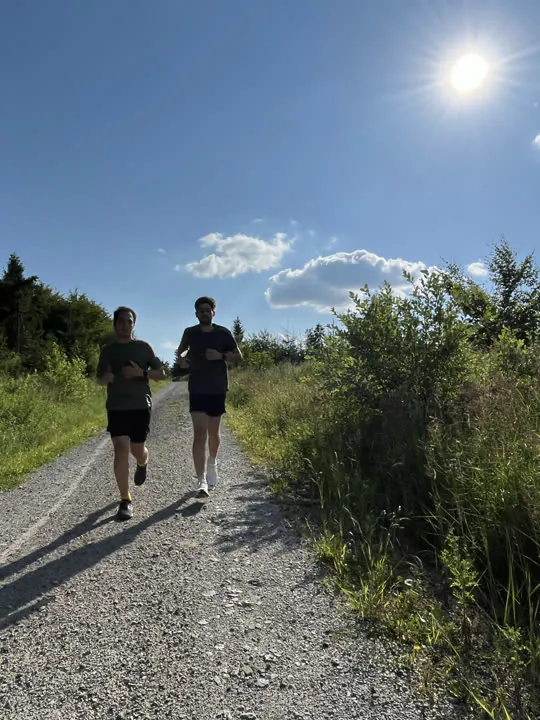 Zwei Kollegen joggen bei Sonnenschein einen Schotterweg entlang